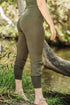 A person wearing high waisted, olive green jogger style leggings standing near a body of water and greenery.