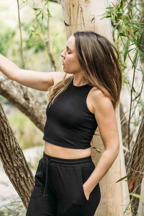 A woman wearing a black bamboo tank top and black pants, standing in a forest setting.