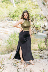 A person wearing a black maxi skirt with a green sleeveless top, standing in a natural setting with trees and water in the background.