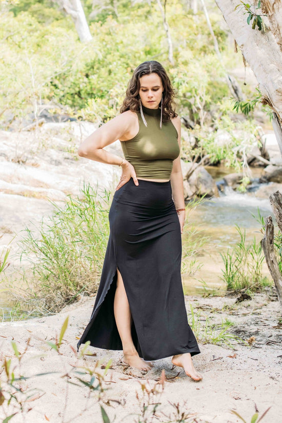 A person wearing a black maxi skirt with a green sleeveless top, standing in a natural setting with trees and water in the background.