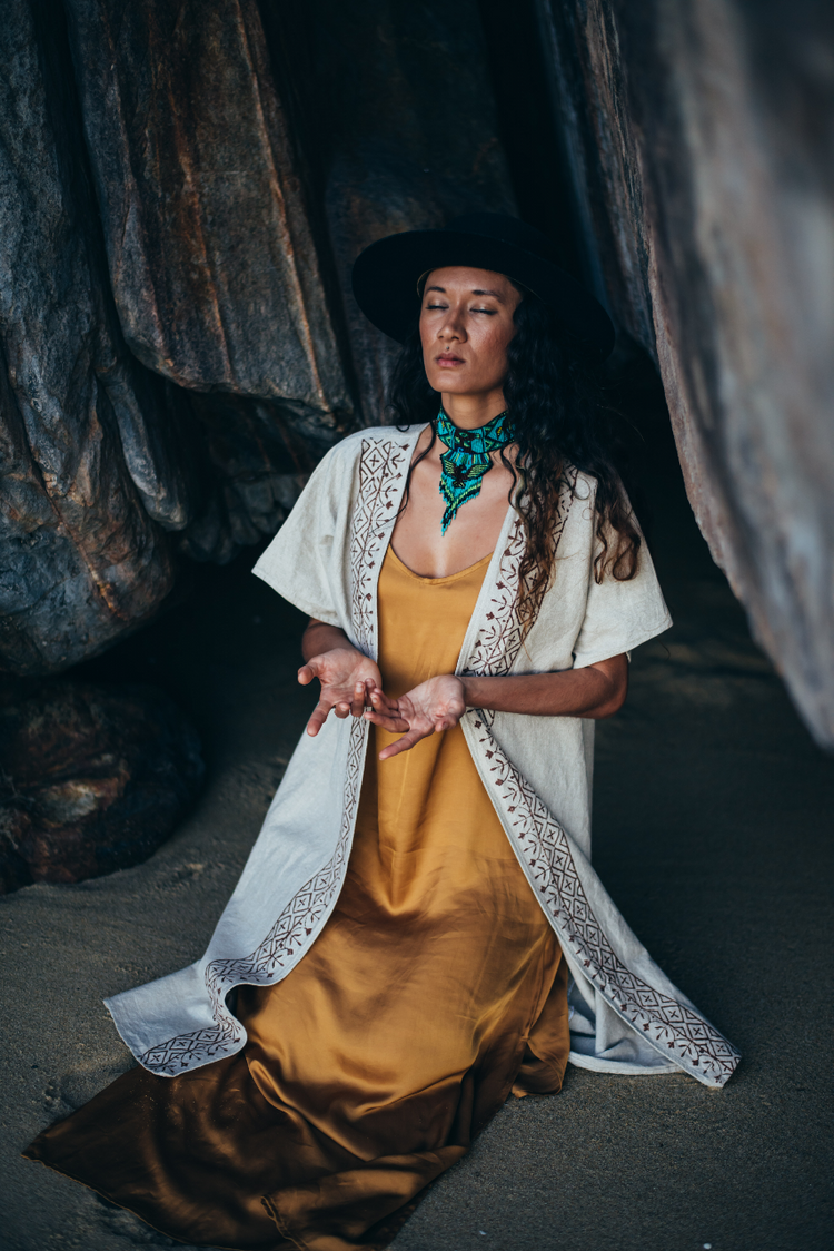 A person wearing a traditional Kaftan-style coat with hand-stamped patterns, paired with a yellow dress and a wide-brimmed hat, standing in a rocky, outdoor setting.