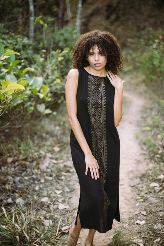 Woman in a black dress with intricate patterns standing on a forest path.