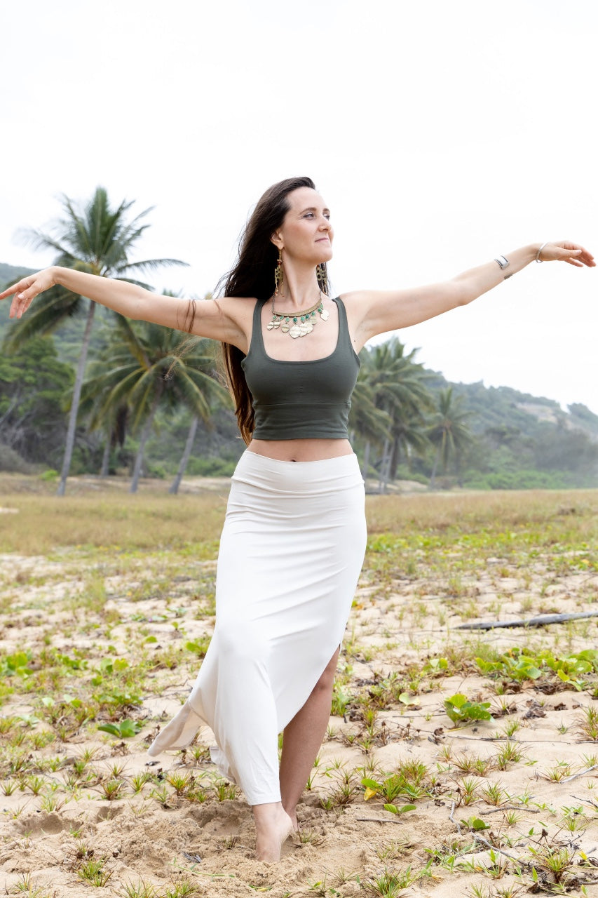 A woman in a green tank top and a white maxi skirt with a double-split design, standing on a sandy beach with palm trees in the background.