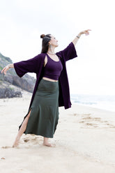 A person standing on a beach wearing a purple kaftan with a flowing skirt and a cropped top, posing with one hand extended gracefully.