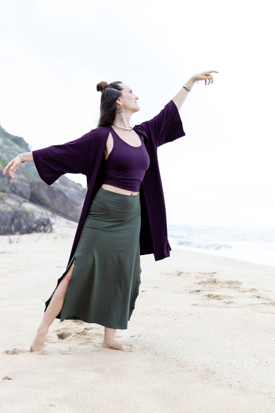 A person standing on a beach wearing a purple kaftan with a flowing skirt and a cropped top, posing with one hand extended gracefully.