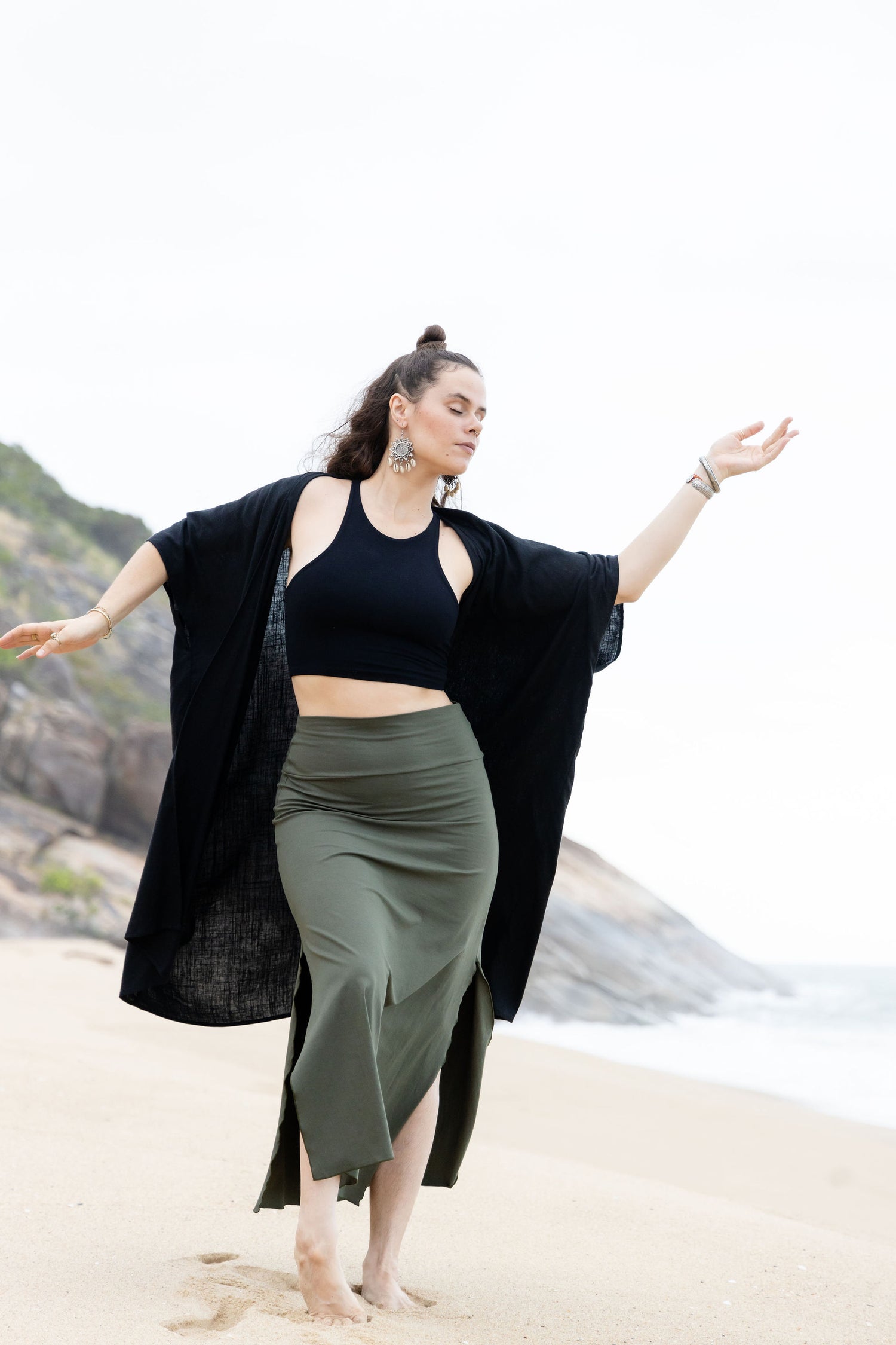 A woman standing on the beach wearing a black Kaftan with long sleeves and a green skirt.