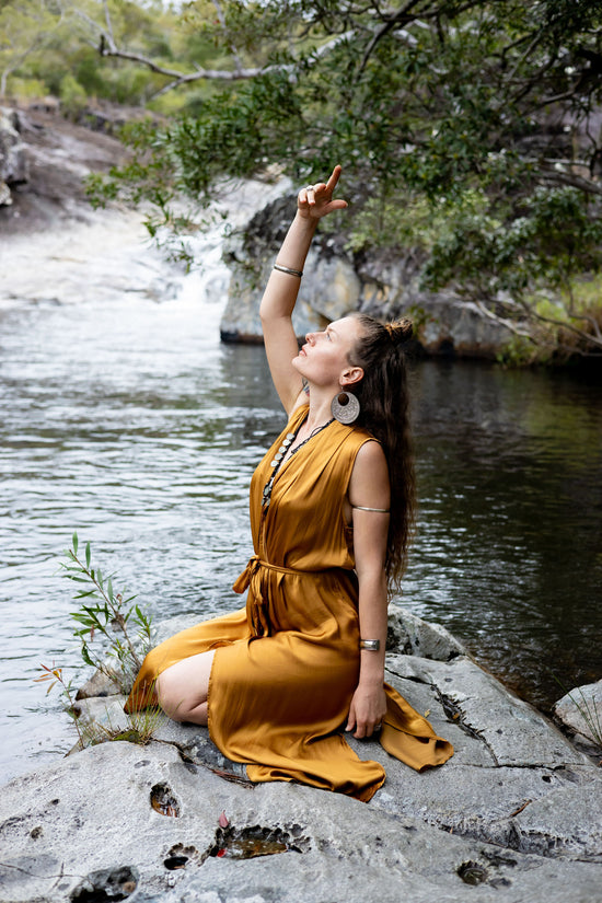A woman sitting on a rock by a riverbank, wearing a copper-colored silk wrap maxi dress with double side splits.